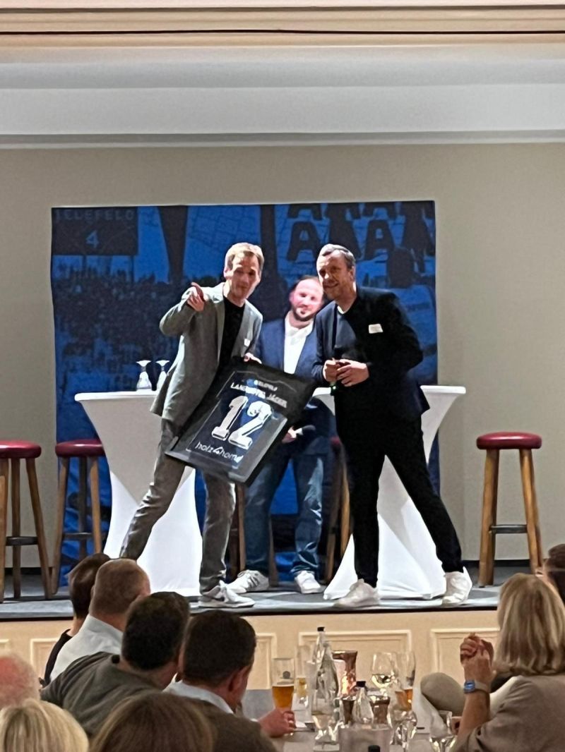 Twee mannen op een podium met ingelijst shirt nummer 12 bij een avondevenement voor sponsors