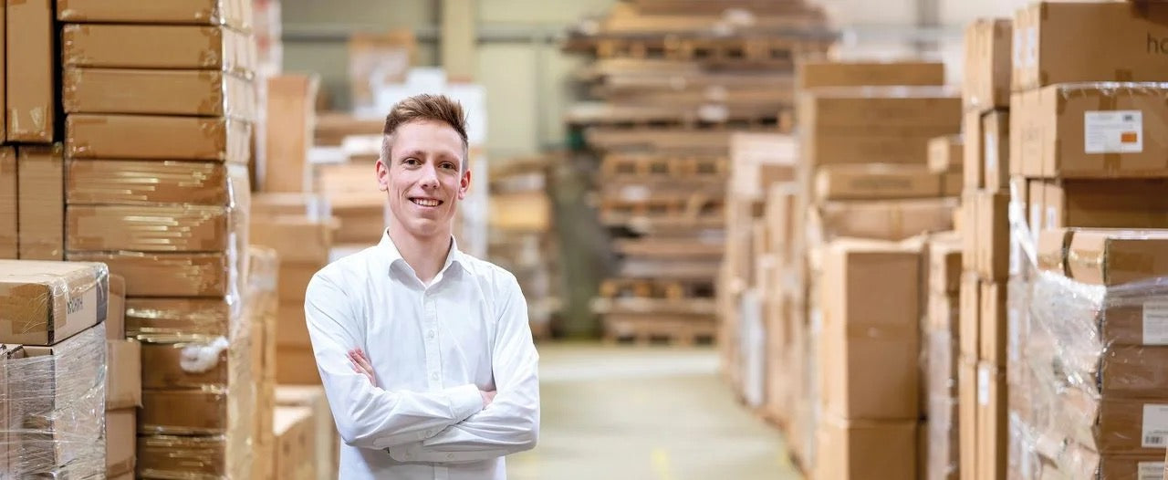 Jonge man in wit overhemd staat met gekruiste armen in een magazijn vol verpakte meubels