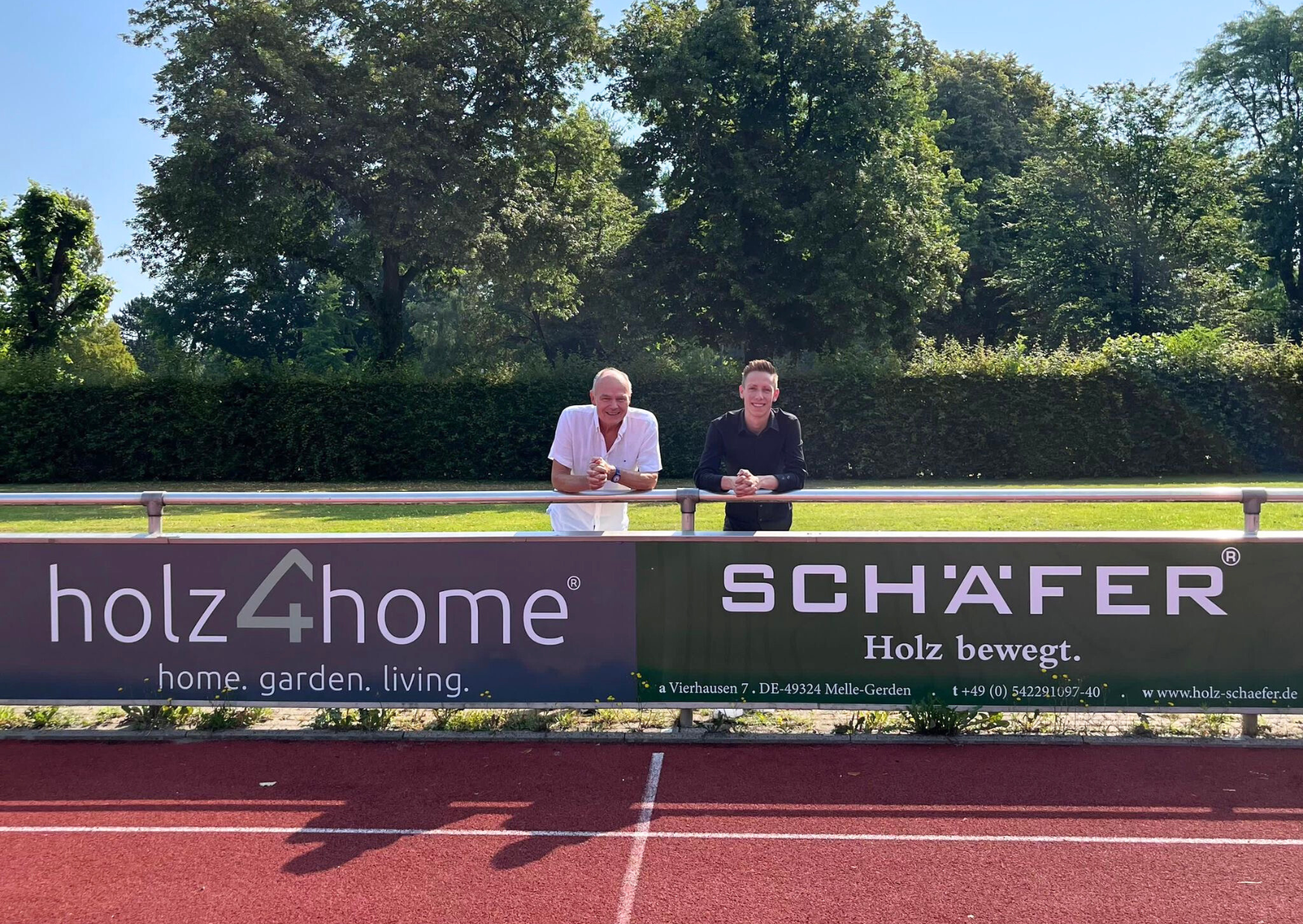 Twee mannen achter een reclamebord van holz4home en Schäfer bij het sportveld van SC Melle 03
