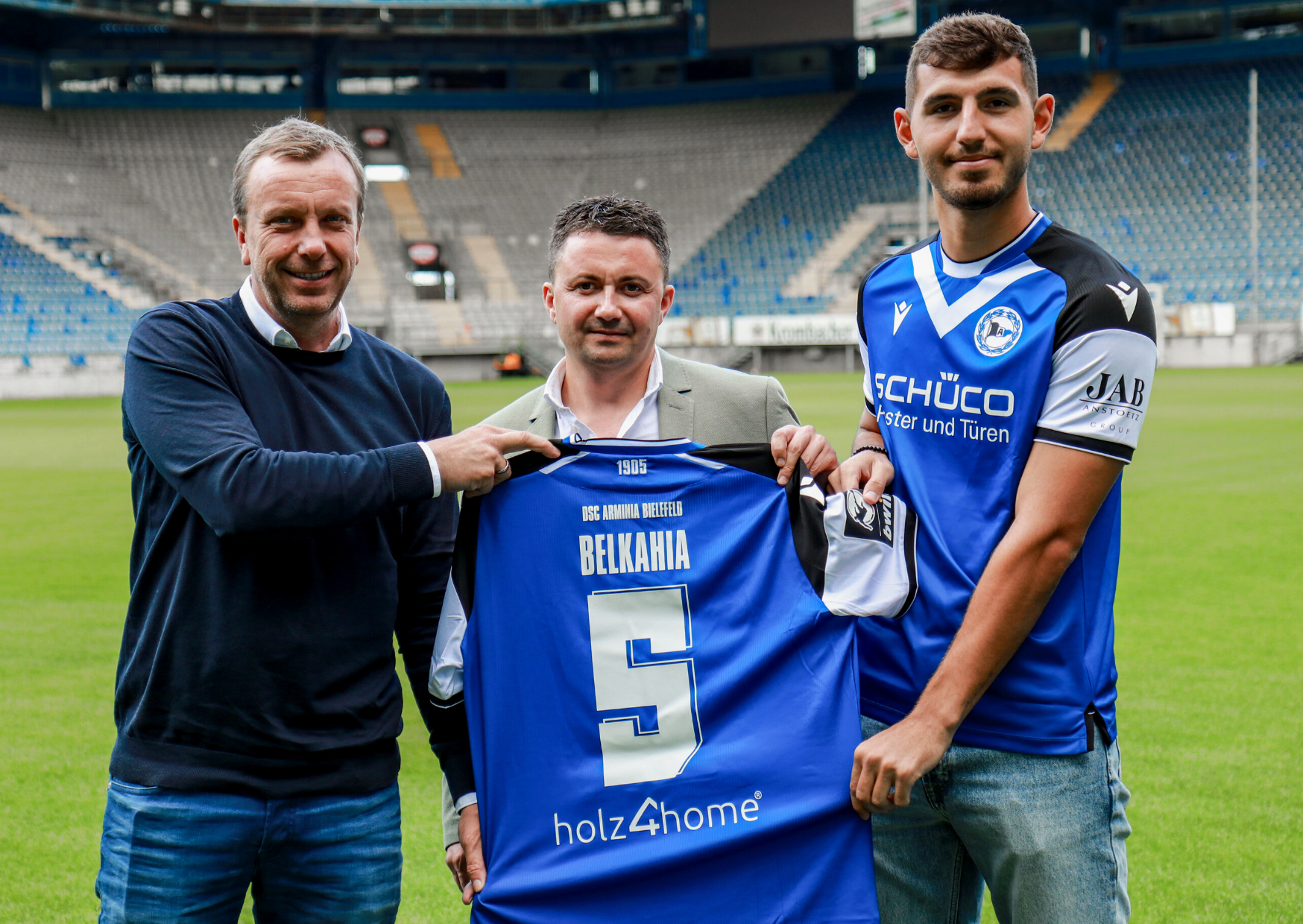 Drei Männer halten ein blaues Trikot von Arminia Bielefeld mit holz4home-Logo im leeren Stadion