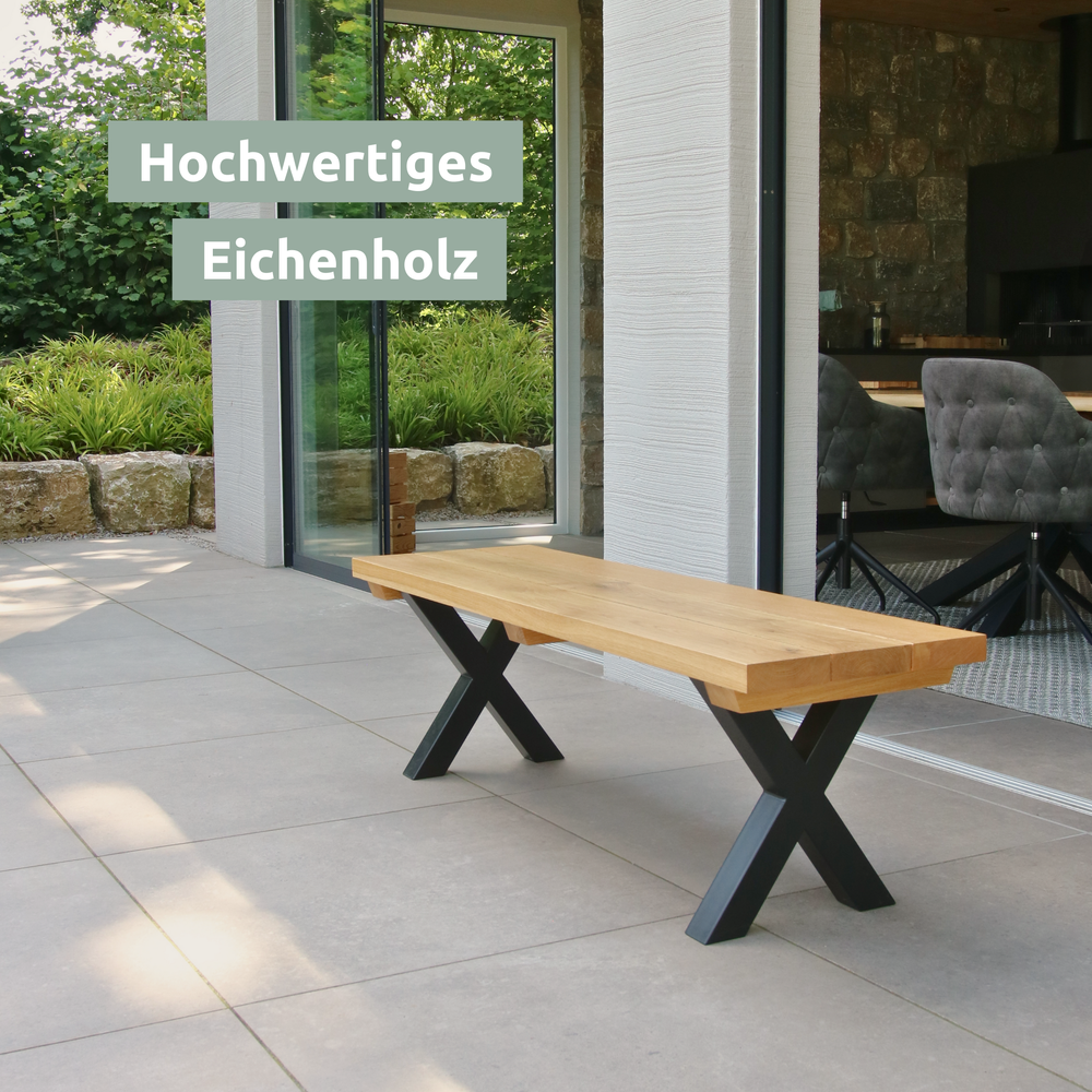 Eiken bank met zwarte X-poten op een terras voor een glazen gevel, met fauteuils op de achtergrond