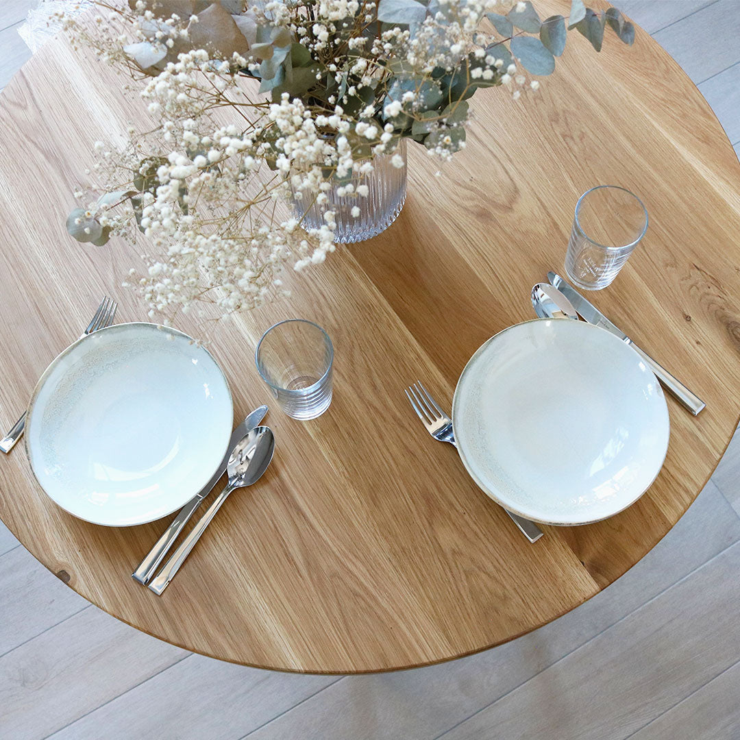 Gedekt rond eikenhouten tafelblad van bovenaf met servies, bestek en droogbloemen